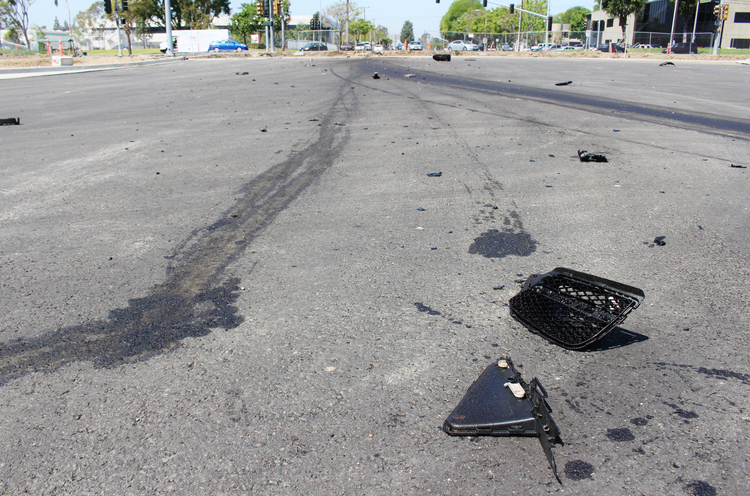 Broken car parts and dark fluid stains are scattered across an asphalt road. The scene suggests the aftermath of a vehicle accident.