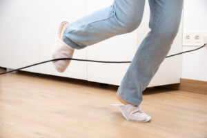A person wearing slippers and jeans trips over a loose electrical cord on the floor. The scene highlights a common household tripping hazard.