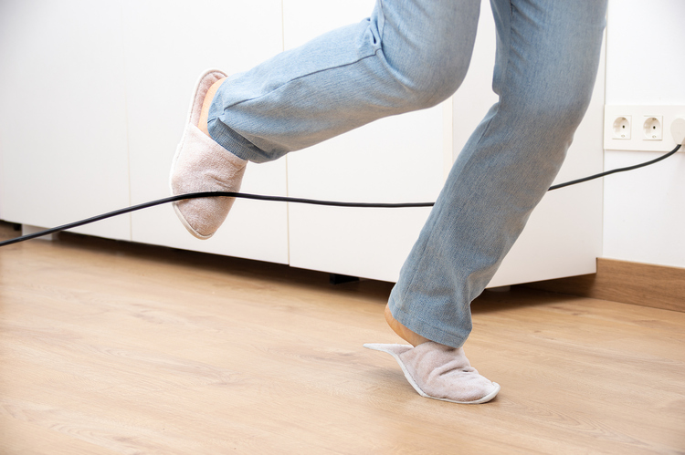 A person wearing slippers and jeans trips over a loose electrical cord on the floor. The scene highlights a common household tripping hazard.