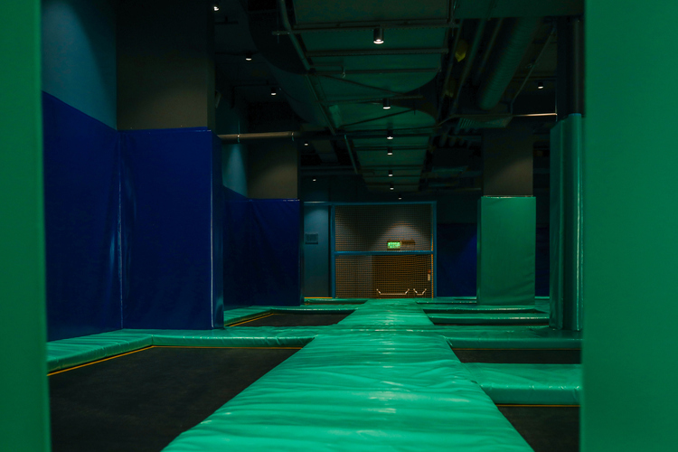 Modern indoor trampoline park with green padded trampolines and support columns. The space is enhanced by netted platforms.